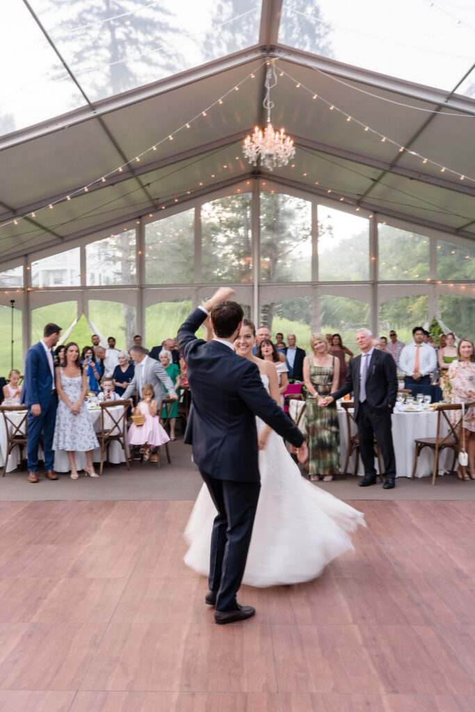 A bride and groom dance together under a clear tent at their Farm Road Estate wedding, surrounded by guests watching and smiling, with tables set for a beautiful reception in the background—capturing timeless Farm Road Estate wedding photos.