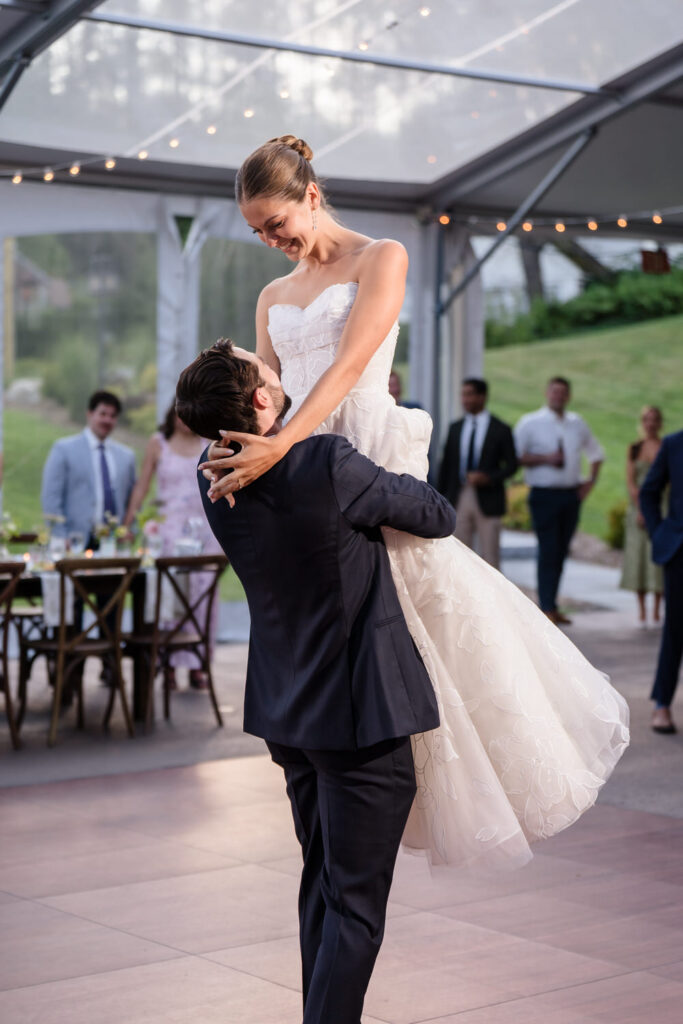 A groom in a dark suit lifts his bride in a white wedding dress while dancing at their outdoor Farm Road Estate Wedding reception, with guests and tables in the background—a beautiful moment captured in these Farm Road Estate Wedding Photos.
