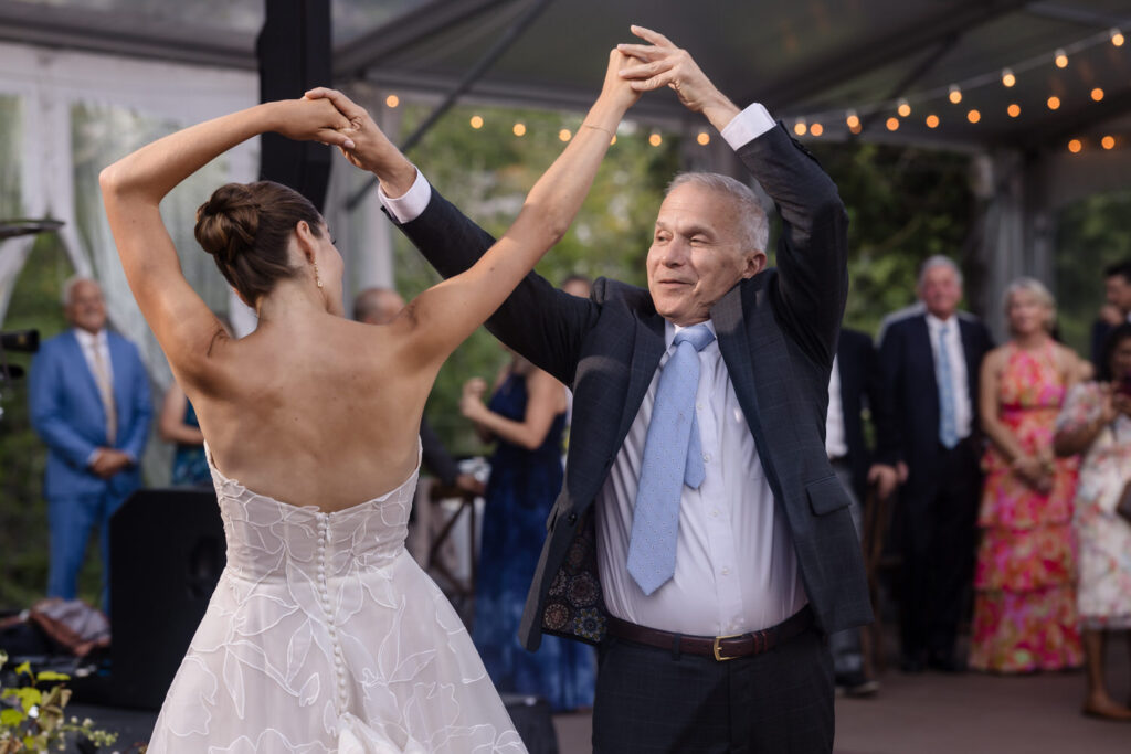 A bride in a white dress dances with an older man in a suit under string lights, surrounded by guests at an outdoor event, beautifully capturing the magic of Farm Road Estate wedding photos.