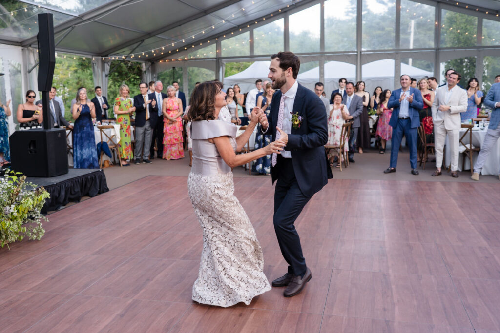 A woman in a lace dress and a man in a suit dance together on a wooden floor as a crowd of well-dressed guests watch and smile inside a tented venue, capturing the elegance and joy reflected in many Farm Road Estate Wedding Photos.