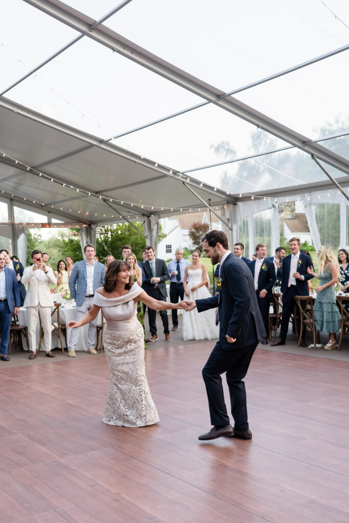 A couple dances joyfully on a wooden floor under a clear tent at Farm Road Estate, surrounded by well-dressed guests watching and smiling at the celebration—capturing the essence of unforgettable Farm Road Estate wedding photos.