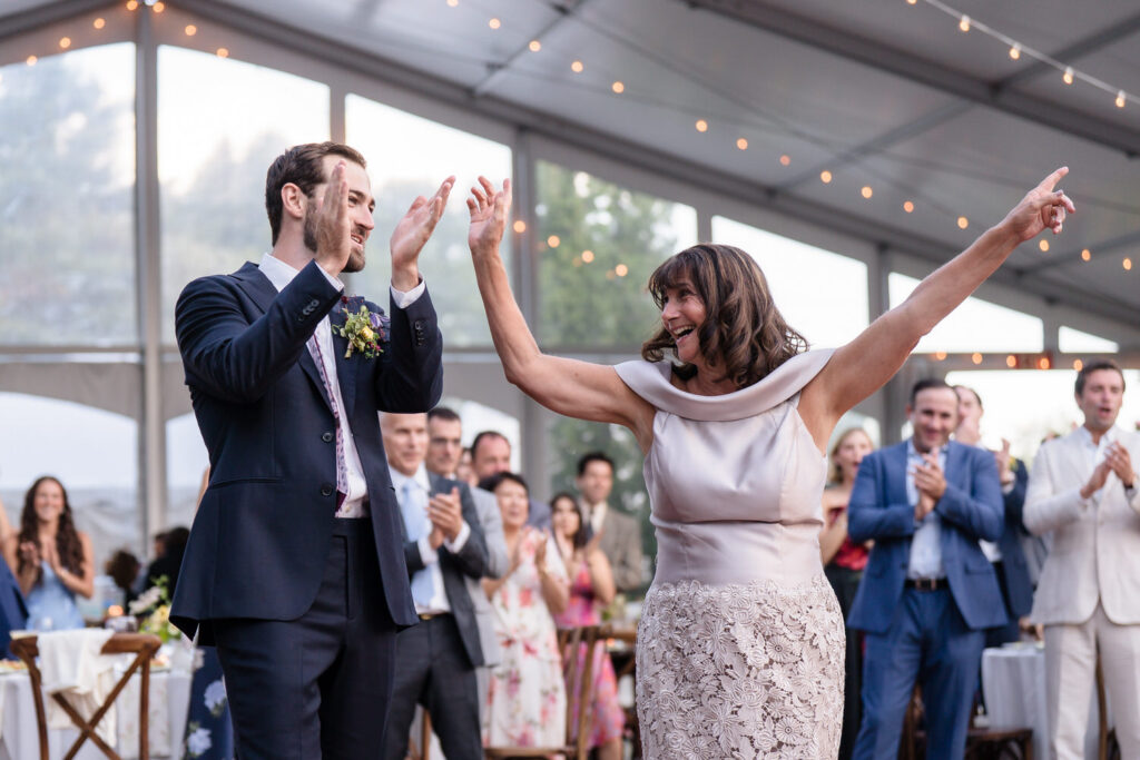 A woman in a light dress dances joyfully with a man in a suit at an event, surrounded by clapping guests under a tent decorated with string lights—capturing the magic of Farm Road Estate Wedding Photos.