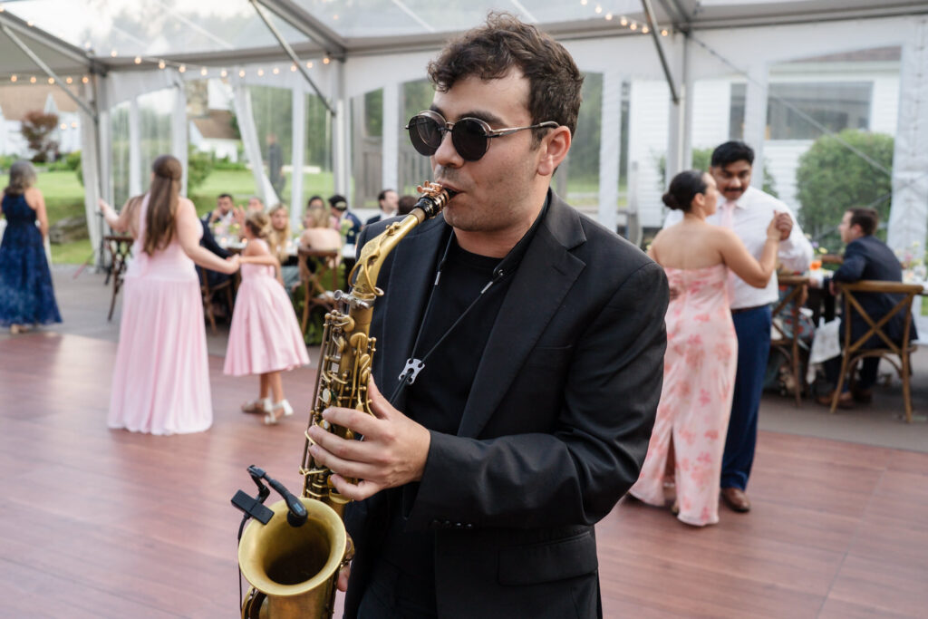 A man in sunglasses plays a saxophone at an outdoor wedding reception at Farm Road Estate, while guests in formal attire dance and socialize in the background under a clear tent.