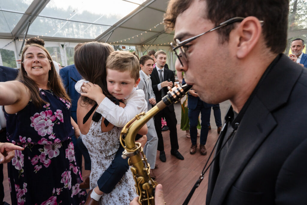 A saxophonist plays at a lively indoor event at the Farm Road Estate, while a woman in a floral dress takes a selfie and a young boy hugs a woman in a white dress. People in formal attire mingle in the background, capturing candid moments perfect for Farm Road Estate wedding photos.