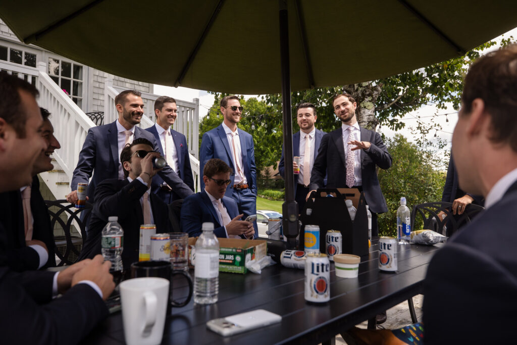 A group of men in suits gather around an outdoor table under an umbrella, laughing and talking—as if sharing memories from a Farm Road Estate wedding. Drinks, snacks, and coffee mugs are on the table. Trees and a white house are visible in the background, evoking the warm ambiance found in candid wedding photos.