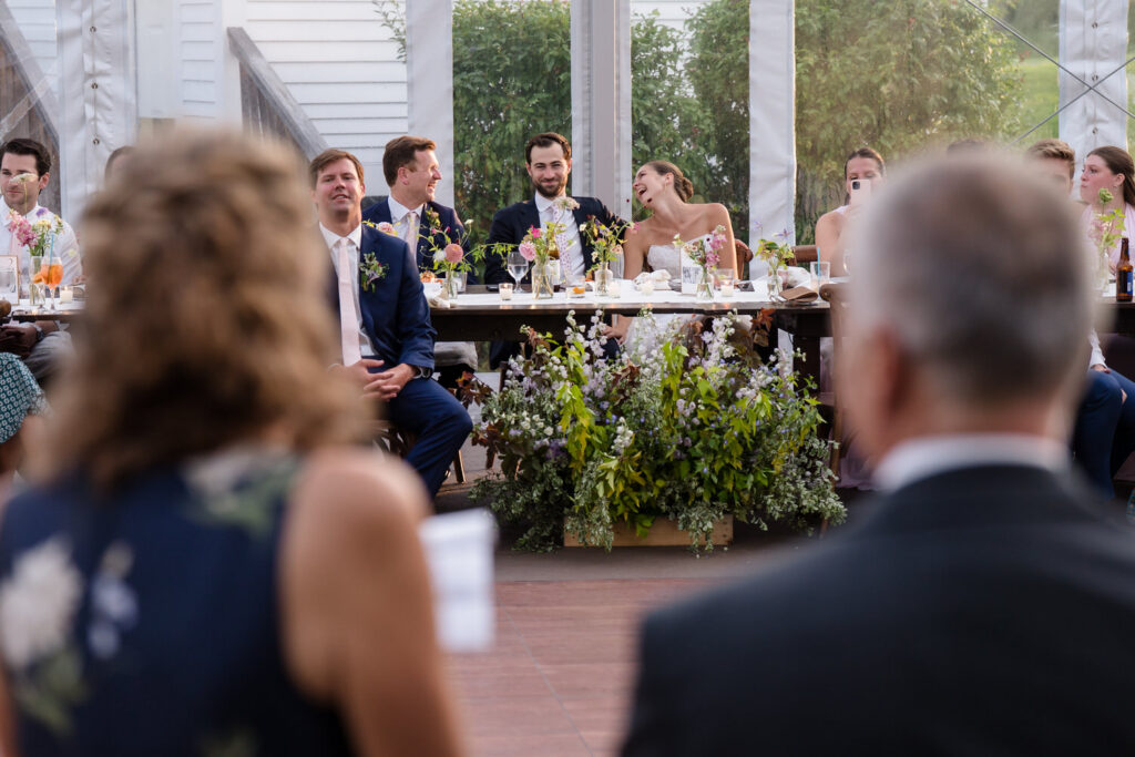 A bride and groom laugh together at a long, decorated table with their wedding party, while two people sit in the foreground, slightly out of focus—a joyful moment captured among beautiful Farm Road Estate Wedding Photos.
