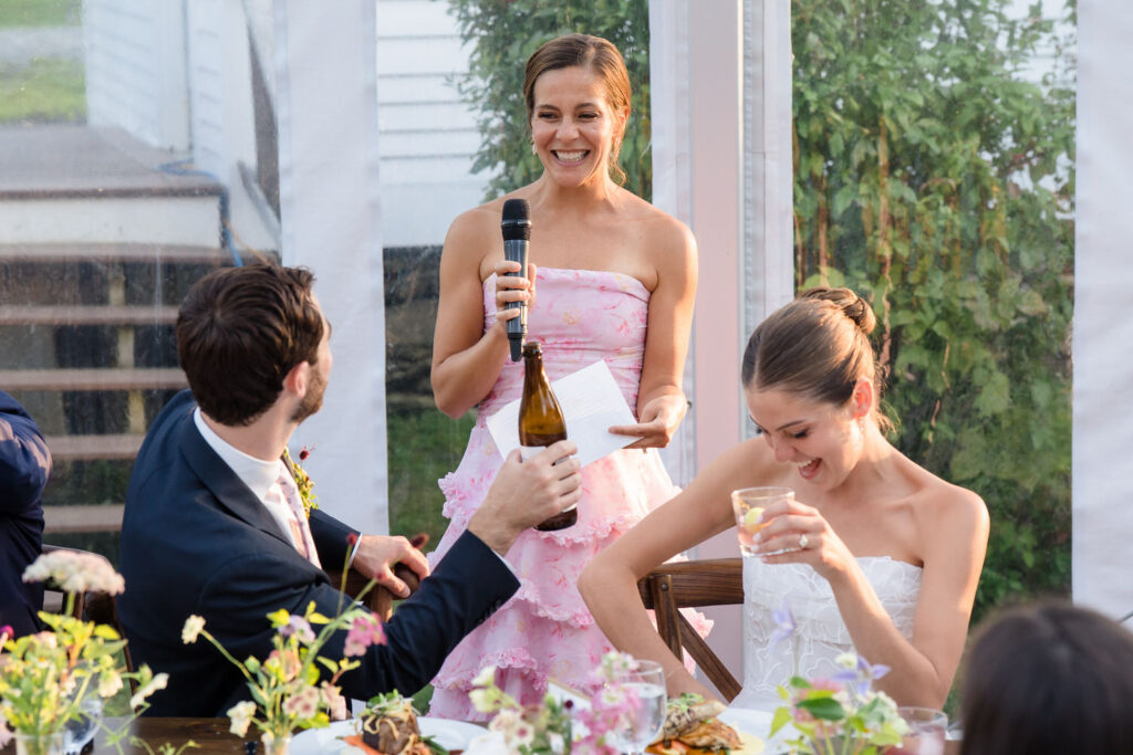 At a Farm Road Estate wedding, a woman in a pink dress gives a speech with a microphone while a man in a suit offers a bottle and the bride, dressed in white, laughs at their beautifully flower-decorated table—capturing joyful moments perfect for Farm Road Estate wedding photos.