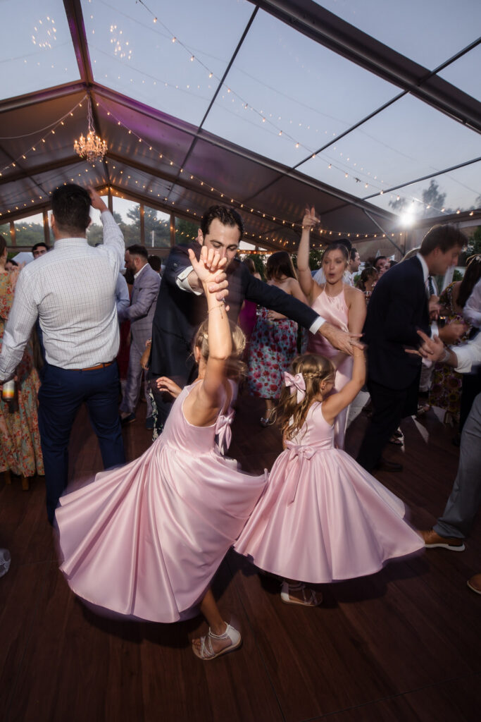 Two young girls in matching pink dresses dance joyfully with a man at a lively Farm Road Estate wedding reception under a clear tent with string lights and a crowd of people celebrating in the background.