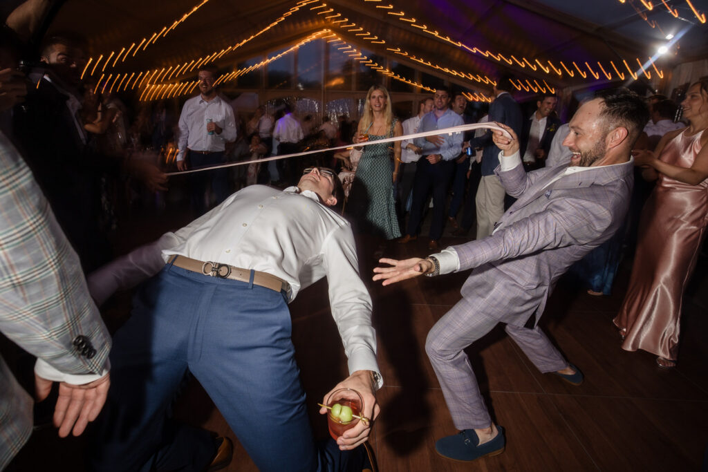 Two men in dress clothes play limbo with a white tie at a lively indoor party while other guests watch and cheer under string lights, capturing the joyful spirit often seen in Farm Road Estate Wedding Photos.
