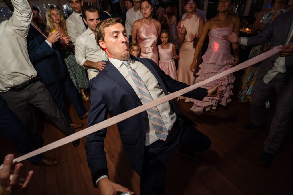 A man in a suit plays limbo at a lively indoor party, bending backward under a ribbon. Guests dressed in formal attire watch and cheer, smiling and enjoying the event—capturing the joyful spirit often seen in Farm Road Estate Wedding Photos.