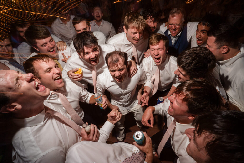 A group of men in white shirts, laughing and singing together in a circle at a lively celebration at Farm Road Estate Wedding Photos, many holding drinks, with festive lighting in the background.