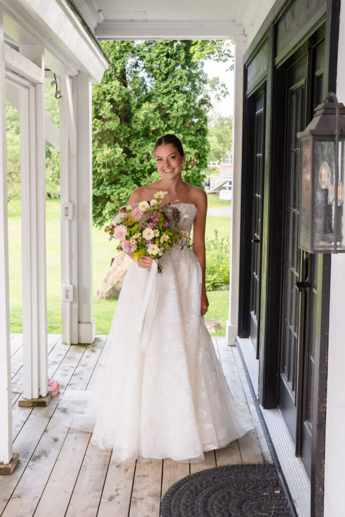 A bride in a white dress stands on a porch at Farm Road Estate, smiling and holding a colorful bouquet of flowers. Green trees and a lawn are visible in the background, perfectly capturing beautiful Farm Road Estate wedding photos.