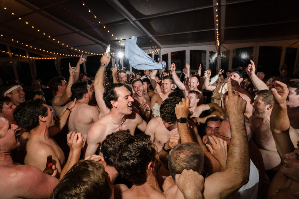 A large group of shirtless men cheer and celebrate together indoors under string lights, raising their arms and appearing joyful in a crowded, lively atmosphere—a perfect example of the energy captured in Farm Road Estate wedding photos.