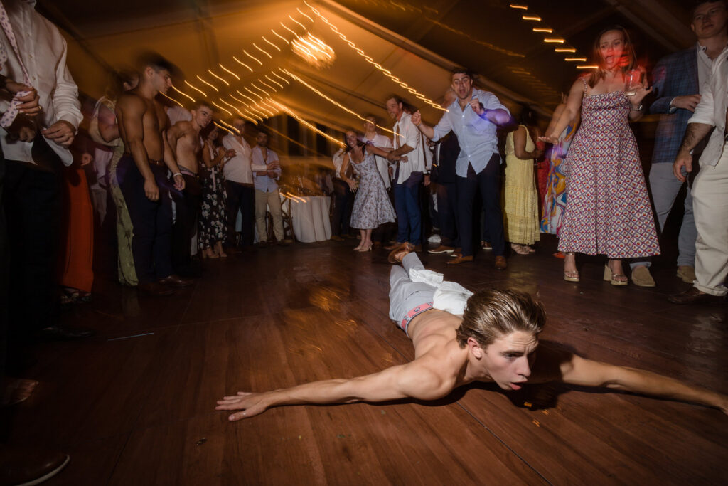A shirtless person slides on their stomach across a dance floor while a crowd of people in formal attire watch and cheer under string lights at a lively Farm Road Estate Wedding.