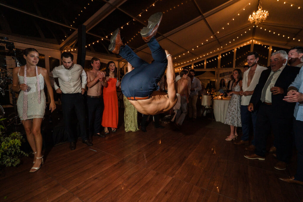 A shirtless man performs a backflip on a wooden dance floor at a party, surrounded by cheering and clapping guests under string lights in a decorated tent—capturing the lively spirit often seen in Farm Road Estate Wedding Photos.