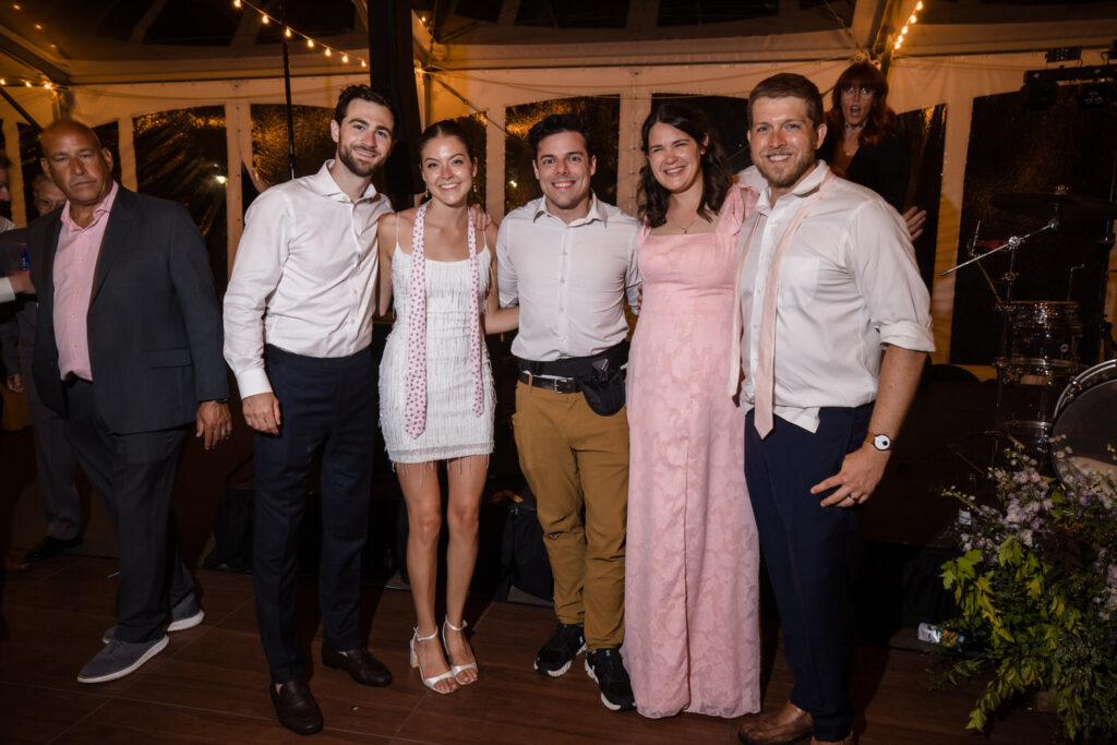 Five people stand together smiling at a party, dressed in semi-formal attire, with string lights and musical equipment in the background—capturing the joyful spirit often seen in Farm Road Estate wedding photos. One person in a pink dress stands out among the group.