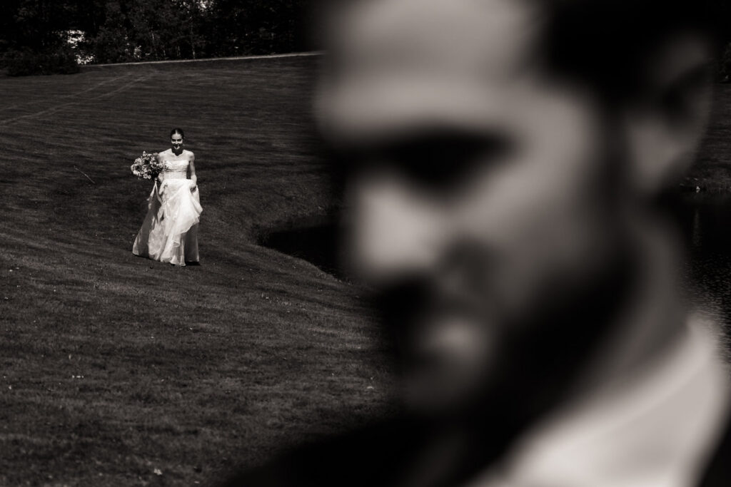 A bride walks across a grassy lawn holding her dress and bouquet, while an out-of-focus man stands in the foreground—a timeless moment captured among the Farm Road Estate Wedding Photos.