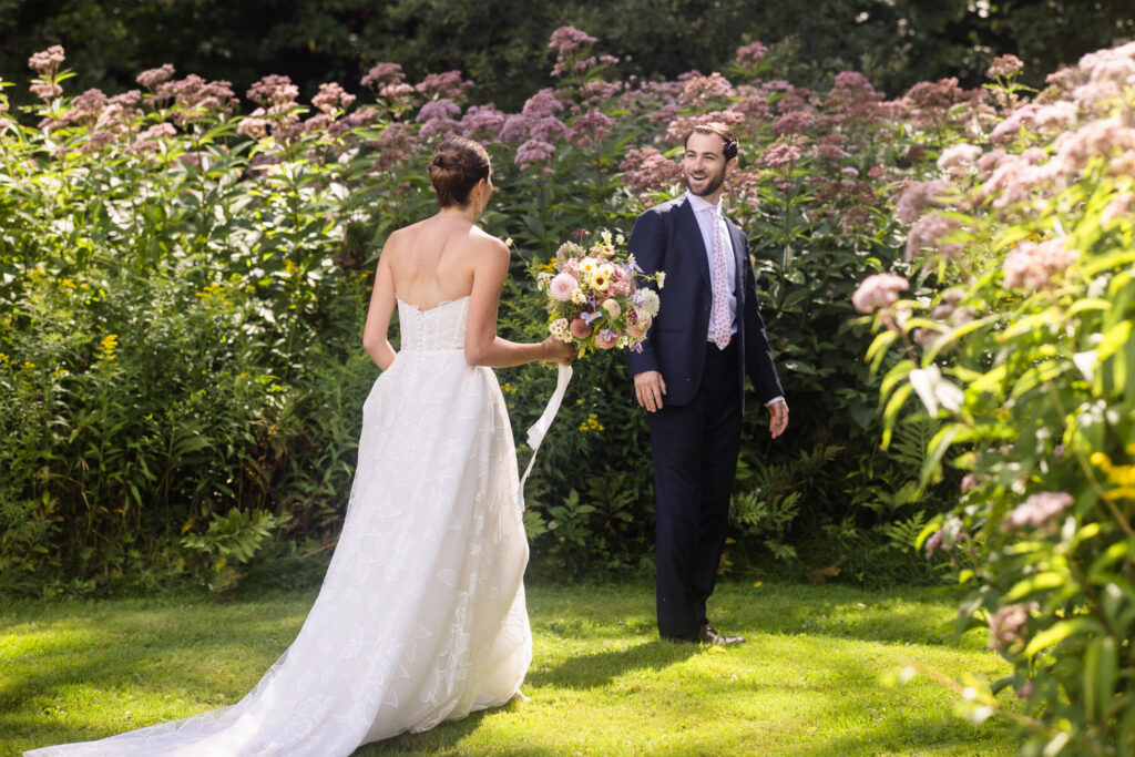 A bride in a white dress walks toward a groom in a suit standing in a garden with blooming flowers, both smiling at each other in the sunlight—capturing the magic of Farm Road Estate wedding photos.