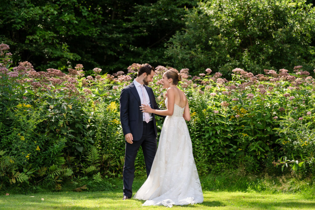 A bride in a white gown and a groom in a suit stand facing each other on grass, surrounded by tall green plants with pink flowers and trees in the background, capturing the natural beauty of Farm Road Estate wedding photos.