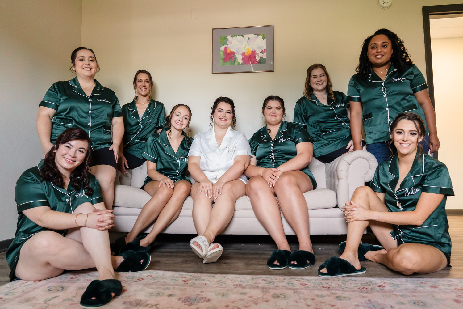 A bride in white pajamas sitting on a sofa surrounded by bridesmaids in matching dark green pajama sets.