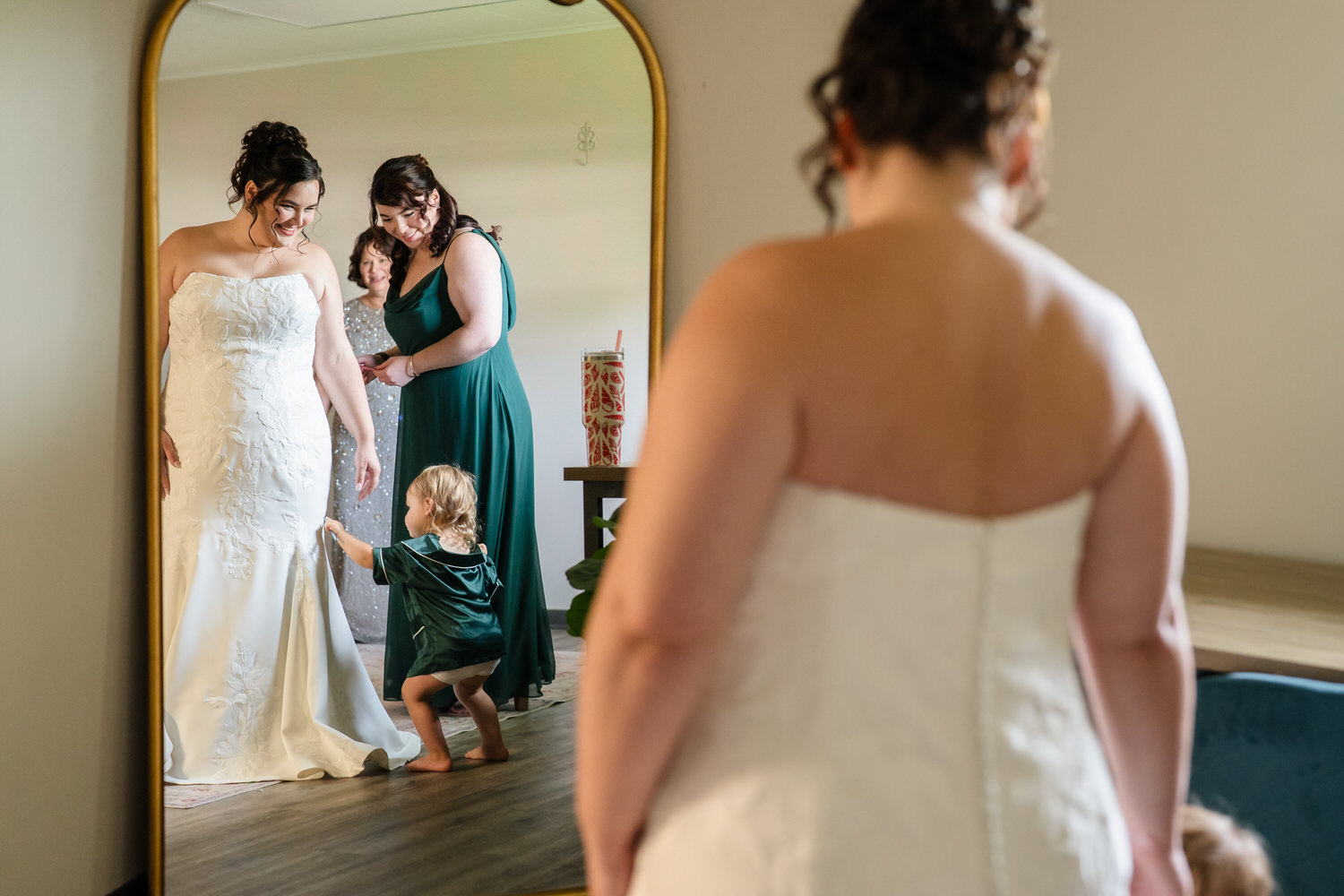 A woman in a wedding dress looks at her reflection in a large mirror, with another woman and a toddler standing beside her.