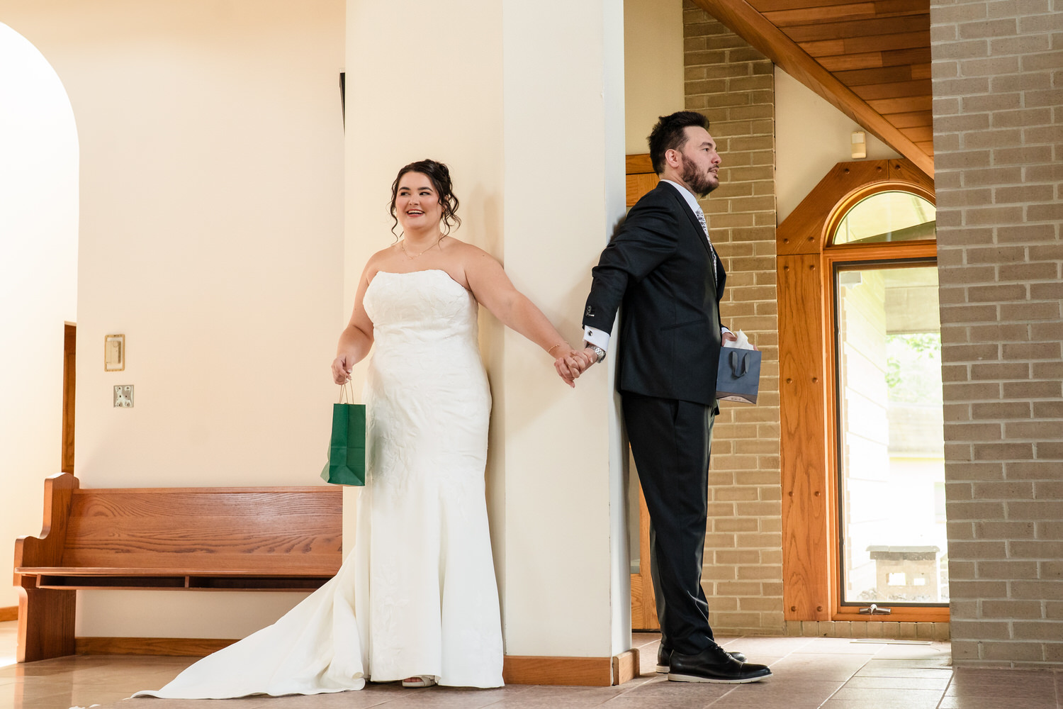 A bride in a white wedding dress and a groom in a black suit stand on opposite sides of a pillar, holding hands without seeing each other.