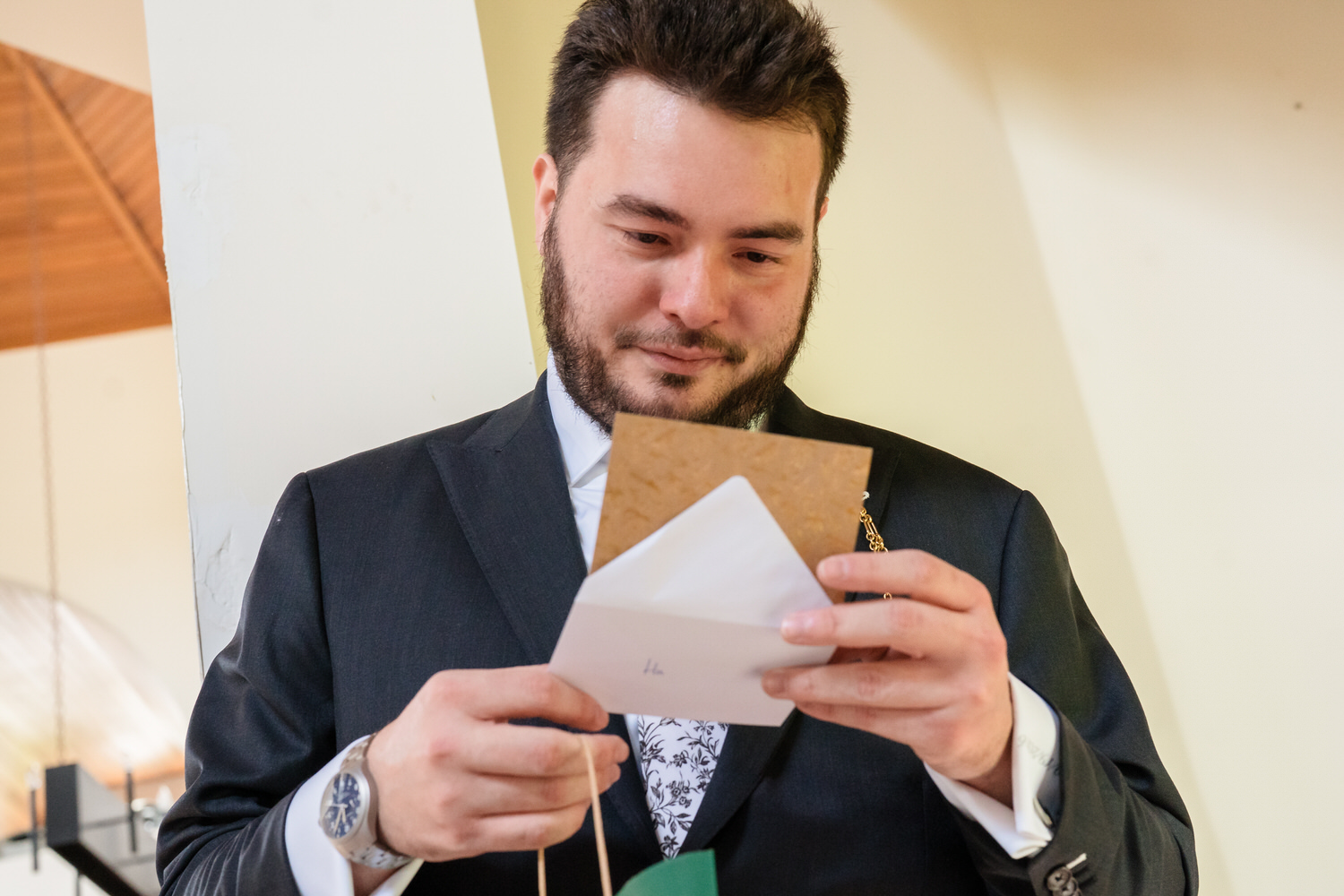 A man with a beard, wearing a black suit and white patterned tie, reads a note from an envelope.