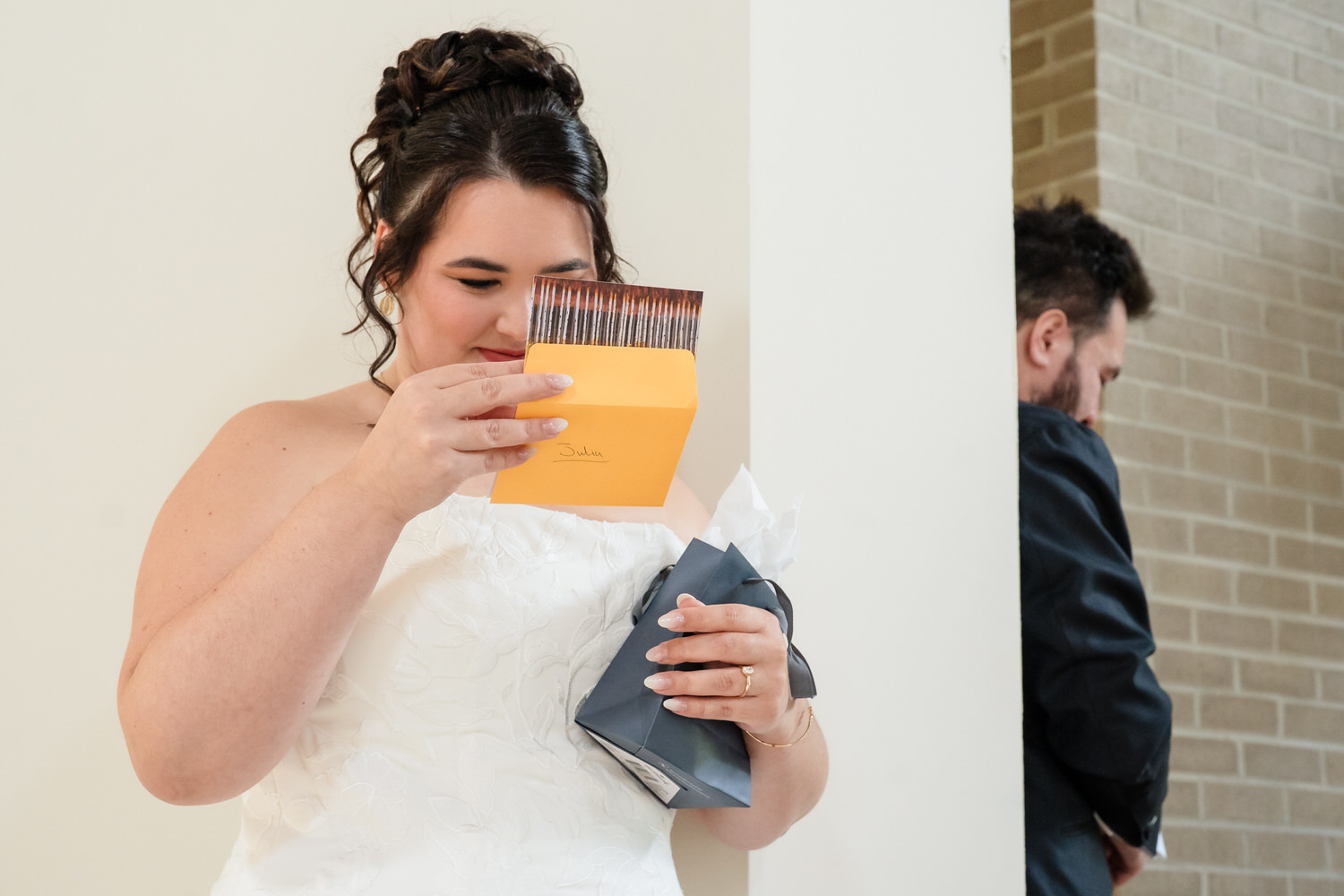 A bride in a white gown opens a gift card marked Julia, while a man stands turned away in the background.