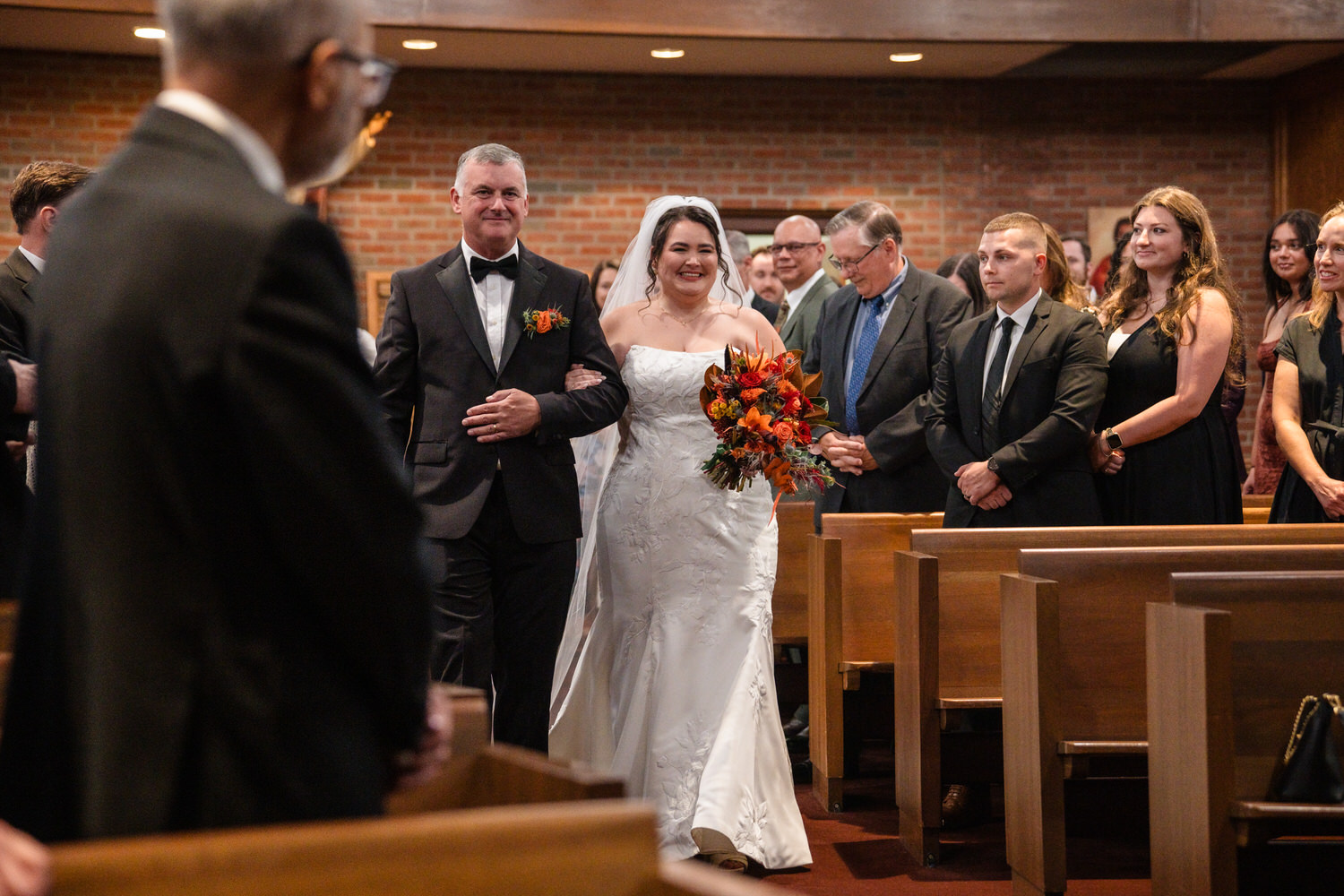 A bride in a white gown and her father walk down the church aisle together as guests watch.