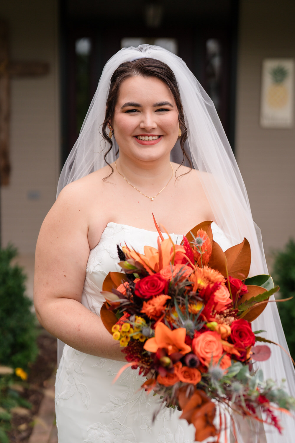 A smiling bride wearing a white wedding dress and veil holds a bouquet of orange, red, and yellow autumn flowers.