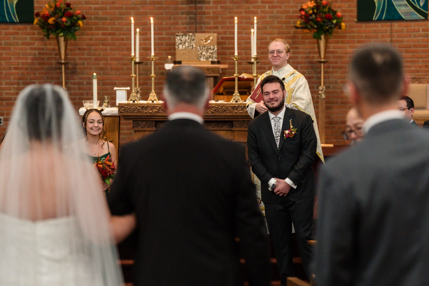 A bride in a white gown and veil walks down the aisle toward a smiling groom in a suit, with an officiant standing behind them at the altar.