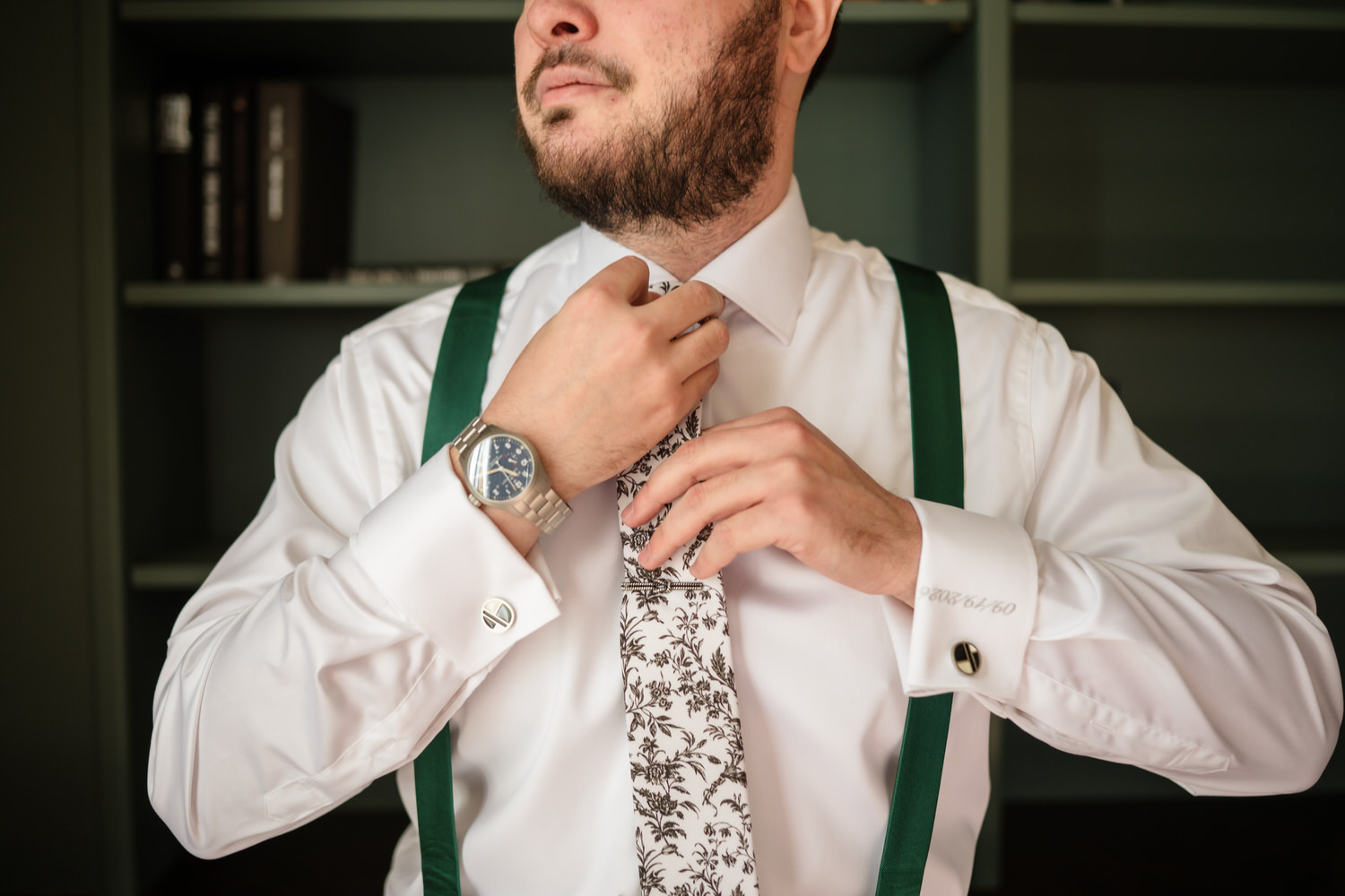 A man in a white dress shirt, green suspenders, and a floral tie adjusts his tie, wearing a wristwatch and cufflinks.