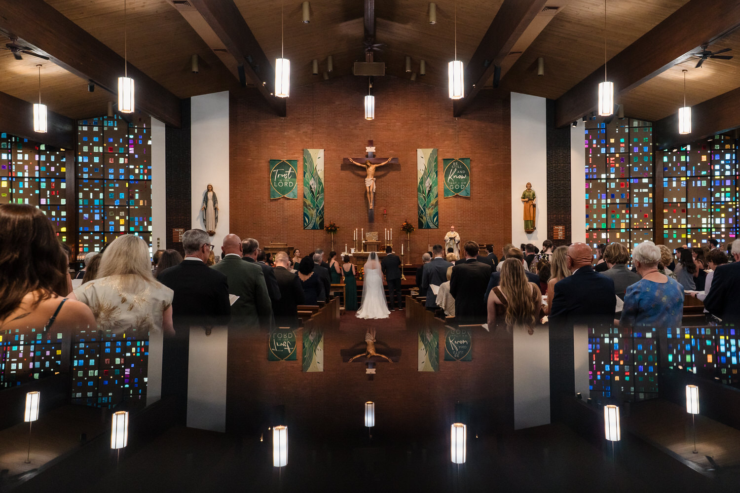 A bride and groom stand at the altar of a church during a wedding ceremony, with guests seated in pews and a reflection of the scene visible in the foreground.