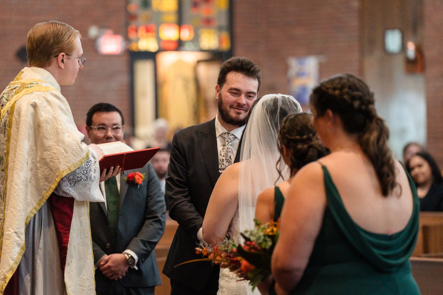 A groom smiles at his bride during their wedding ceremony as an officiant reads from a book.