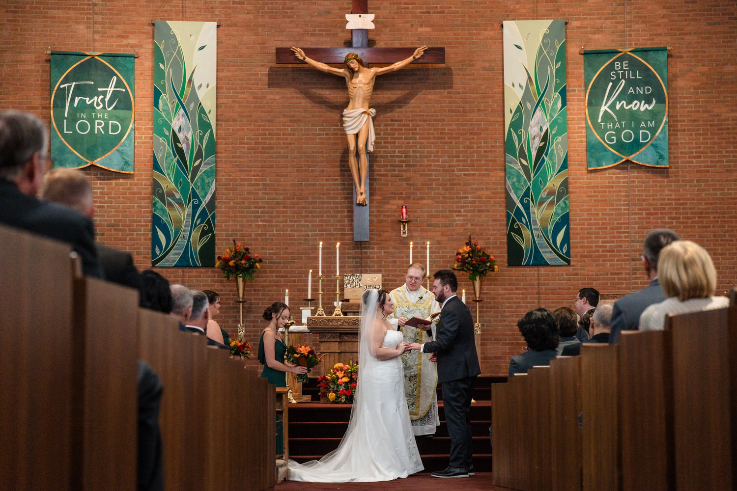 A bride and groom stand at the altar during a wedding ceremony inside a brick church, with a priest officiating before a large crucifix.