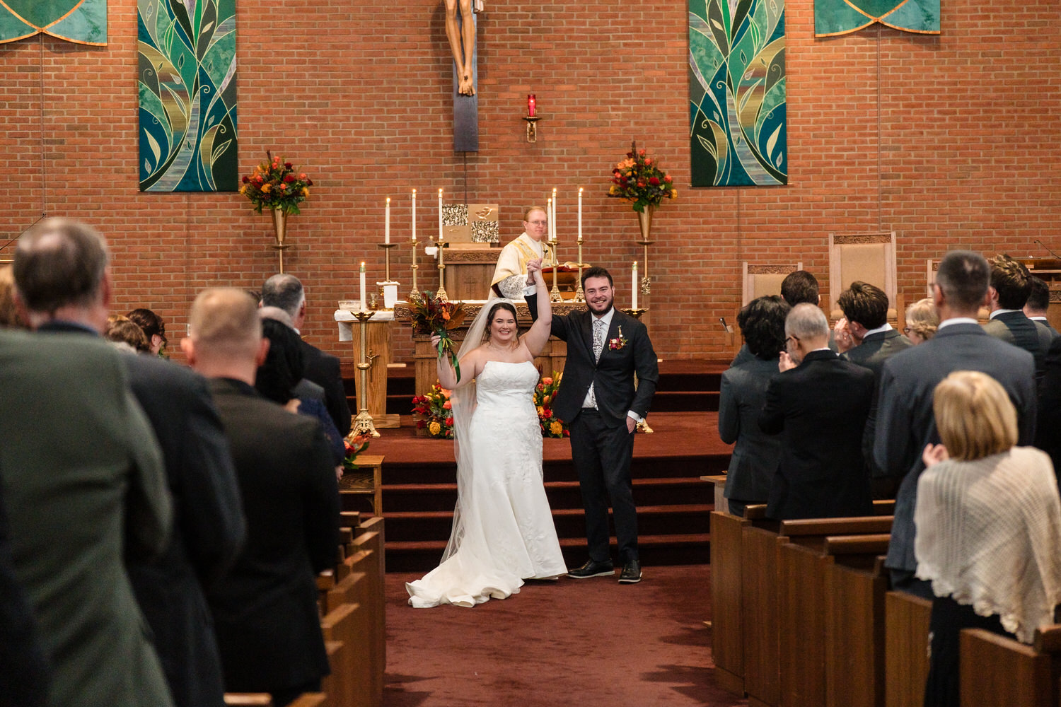 A bride and groom walk down the aisle together after their wedding ceremony in a church, with the groom raising his arm in celebration.