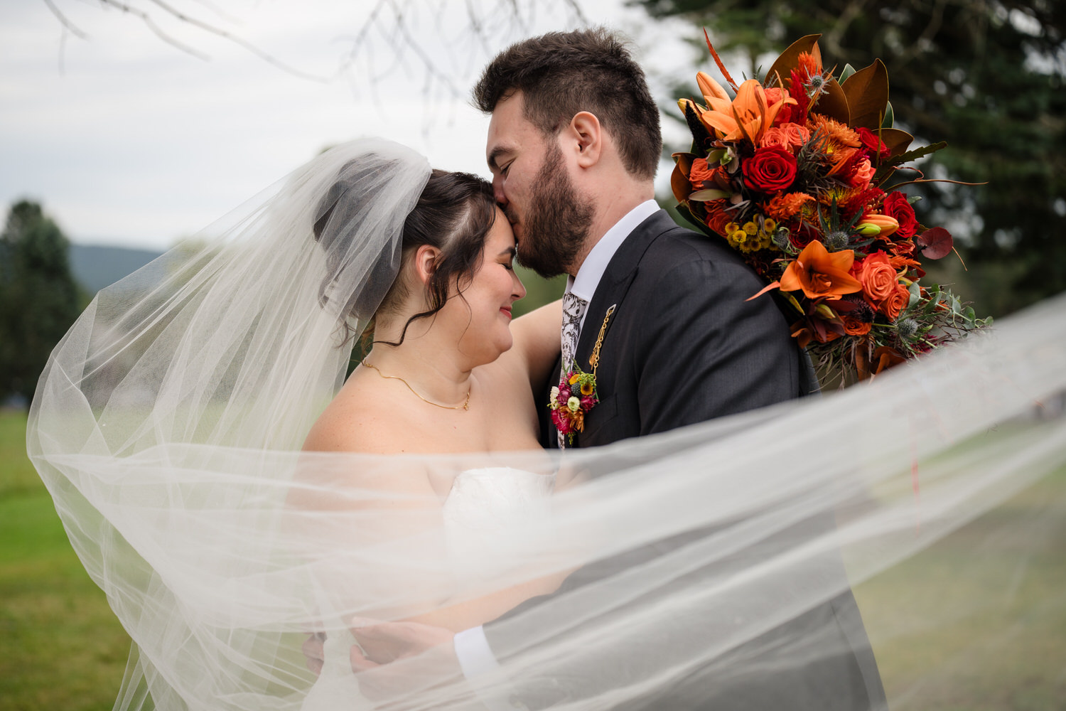 A groom kisses the forehead of his bride, who wears a veil blowing across the foreground, while holding a large bouquet of orange and red flowers.