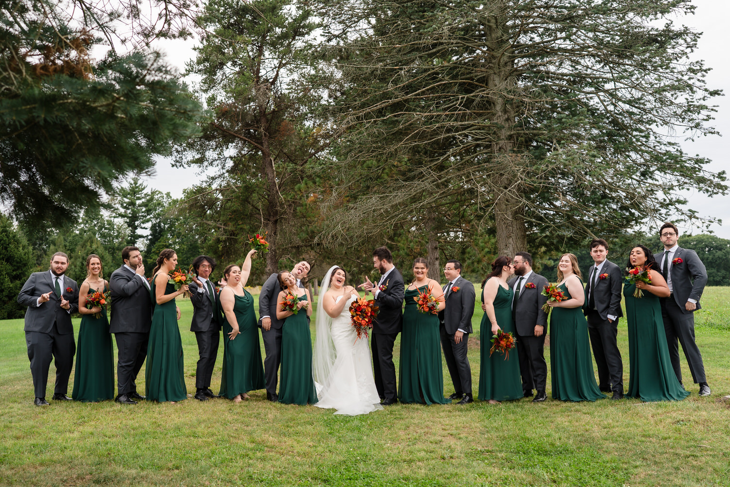 A bride and groom stand in the center of their wedding party in a grassy park, with many of the bridesmaids and groomsmen laughing and posing for the camera.