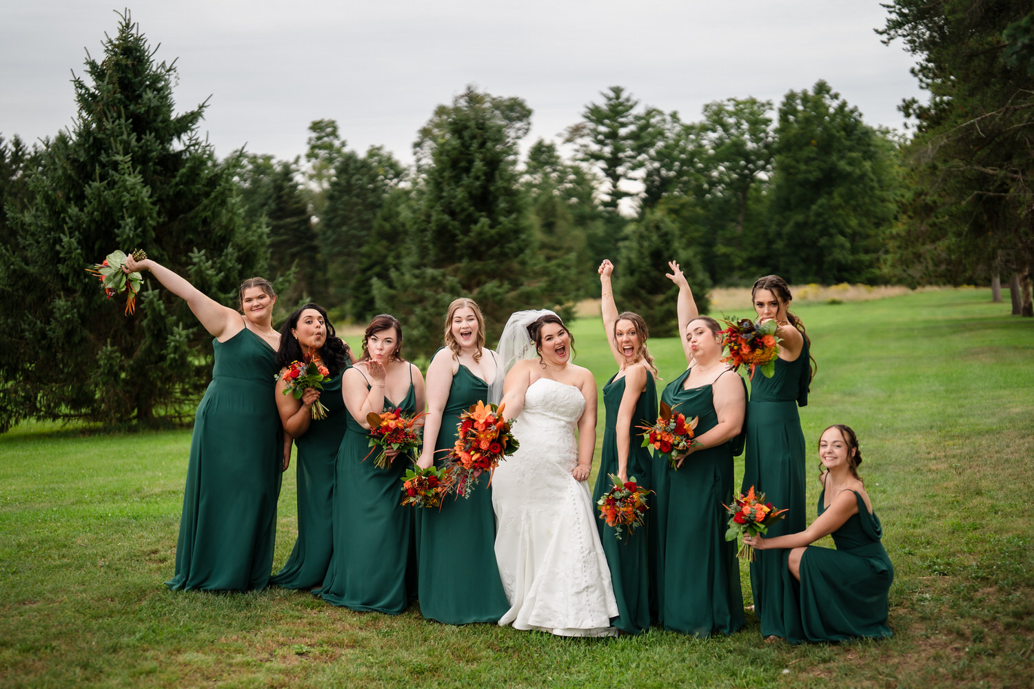 A bride in a white wedding gown posing with her nine bridesmaids in dark green dresses, all holding fall-themed bouquets in a grassy field.