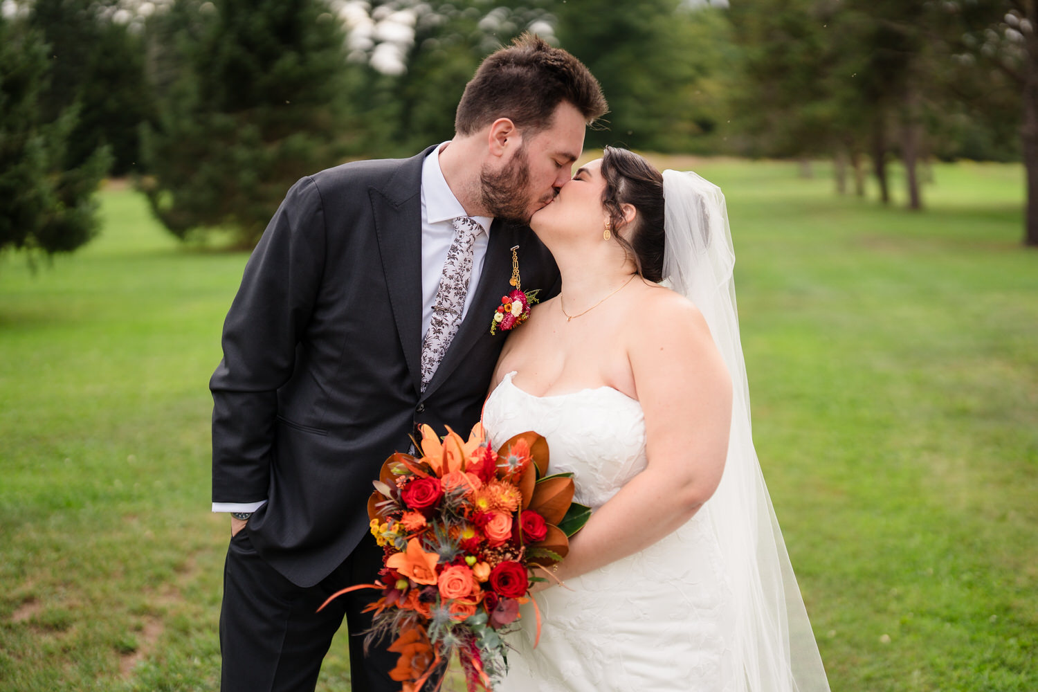 A bride and groom in wedding attire kiss while holding a bouquet of orange, red, and brown flowers in a grassy field.