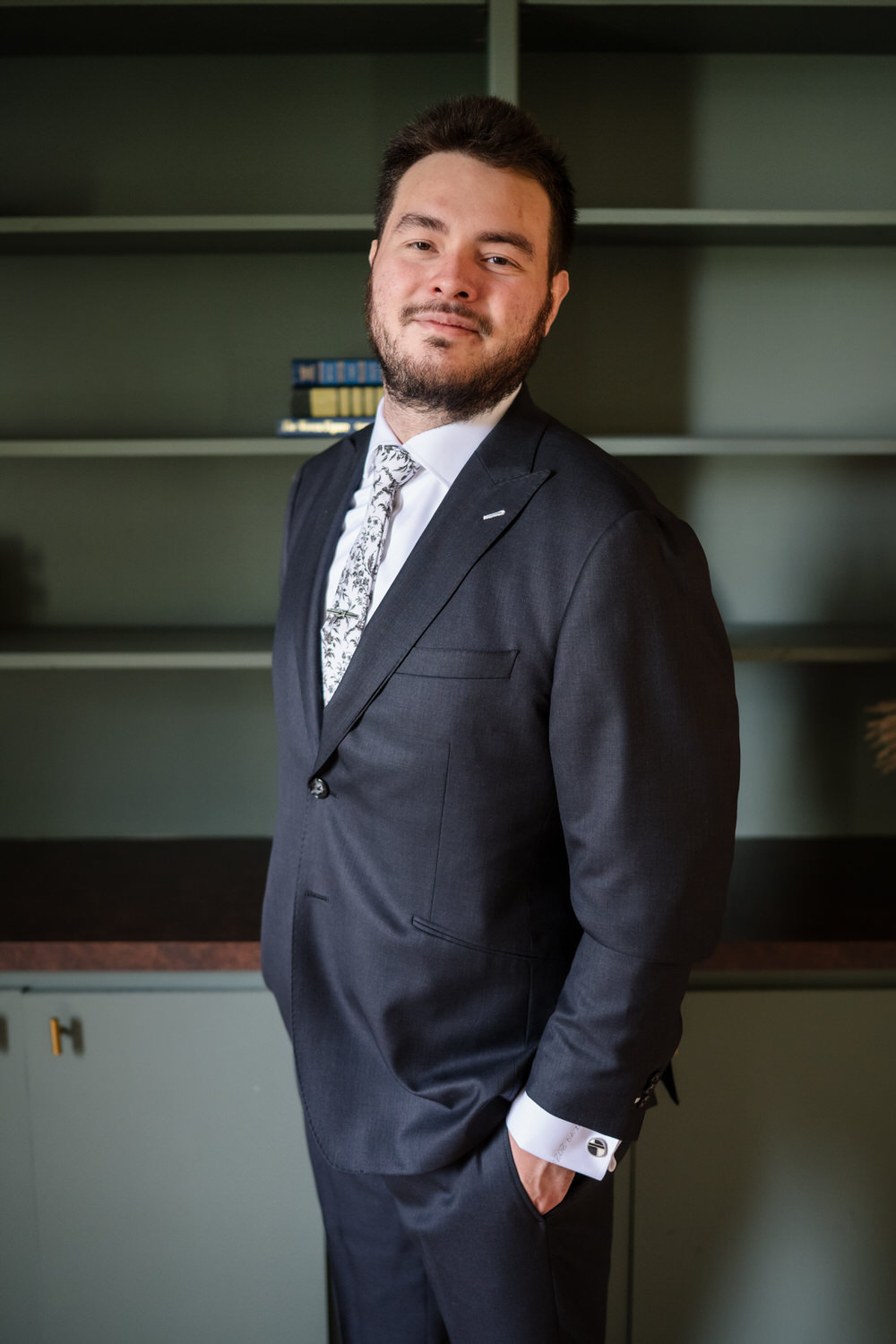 A man with a beard wearing a dark suit and a patterned tie, standing in front of a green bookshelf.