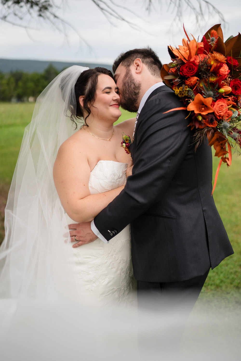 A bride in a white wedding dress and veil stands in a field as the groom, wearing a dark suit and holding a large bouquet of autumn-colored flowers, kisses her cheek.