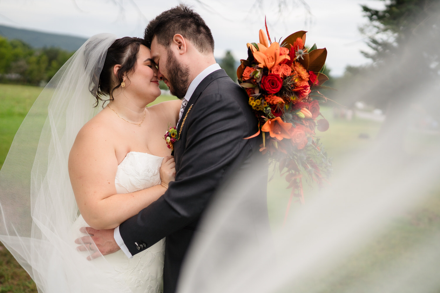 A bride in a white gown and a groom in a dark suit embrace with their foreheads touching, the bride holding a vibrant bouquet of orange and red flowers.