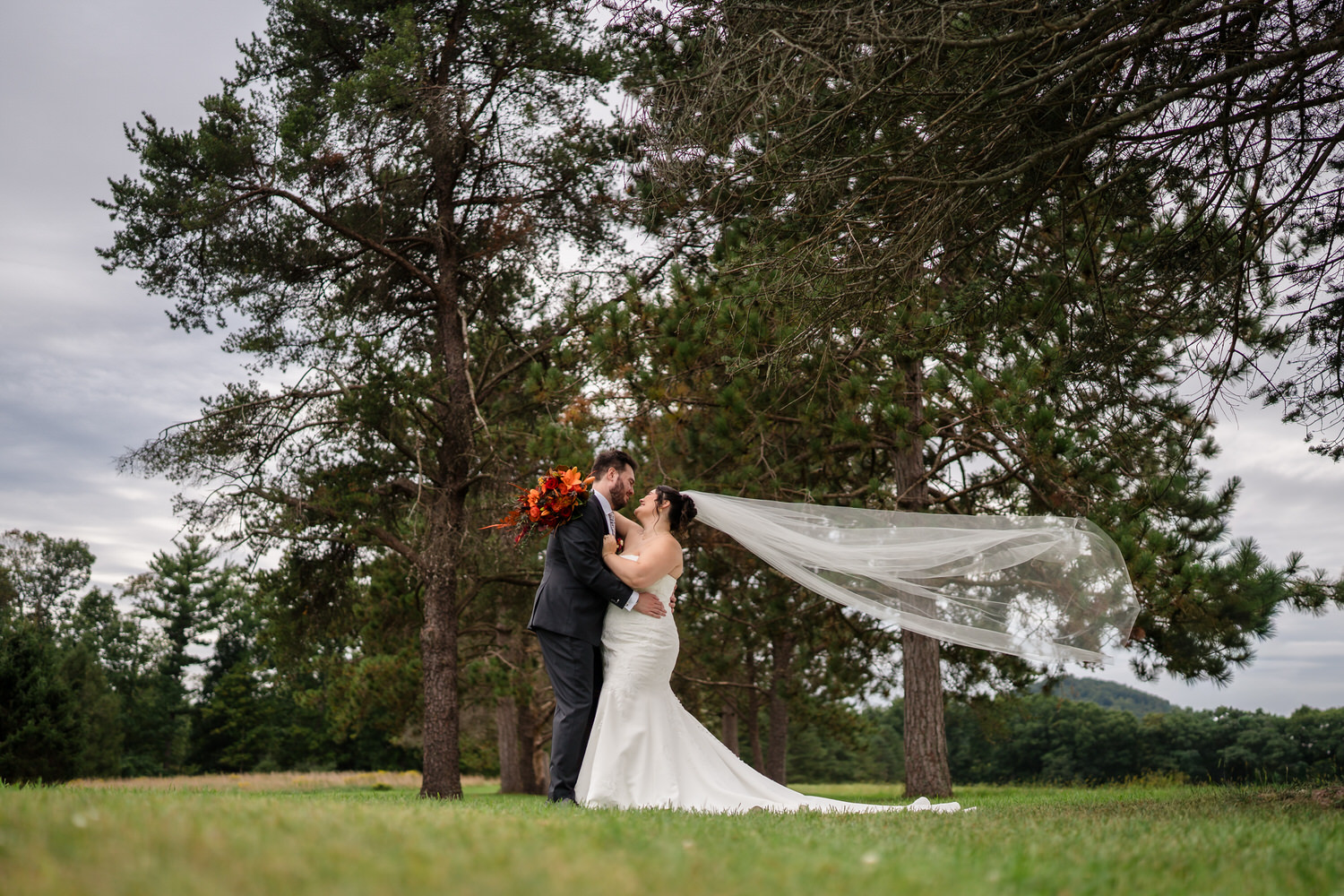 A bride and groom standing in a grassy field with the brides long veil blowing in the wind.