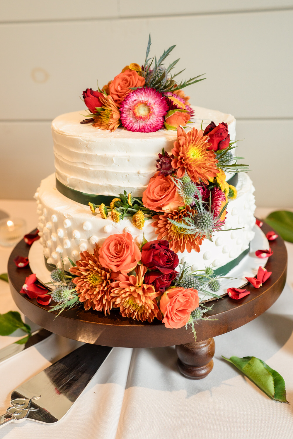A two-tiered white cake on a wooden pedestal, decorated with vibrant orange, red, and yellow flowers and greenery.