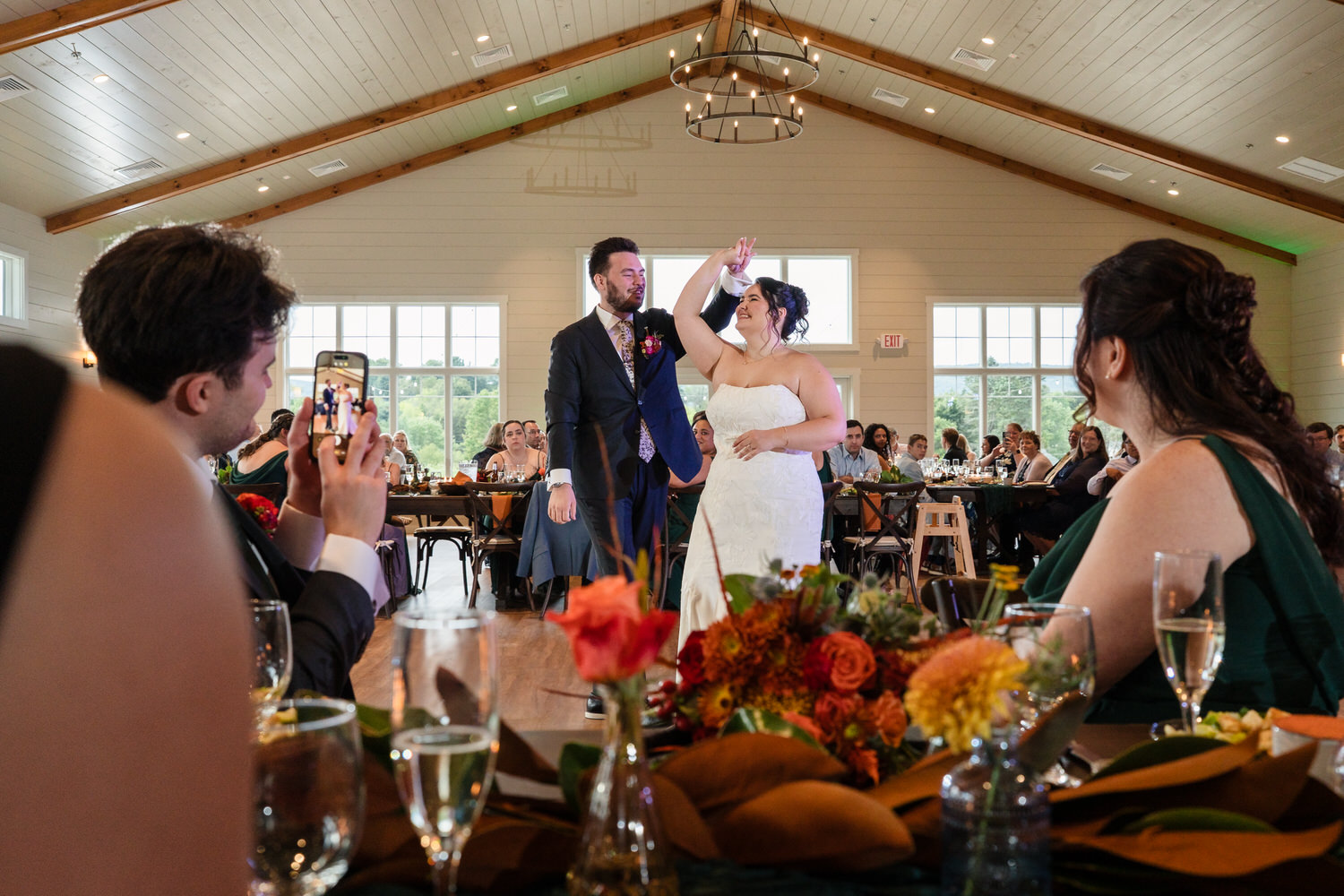 A bride and groom share a dance in the center of a wedding reception hall as guests watch and take photos from their tables.
