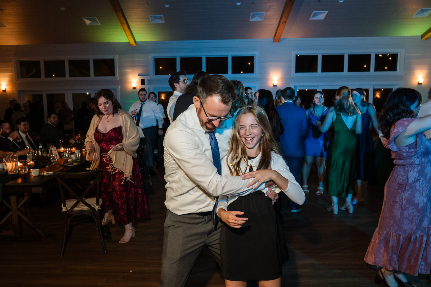 A man in a white dress shirt and tie dancing with a laughing young girl in a black dress at a wedding reception.
