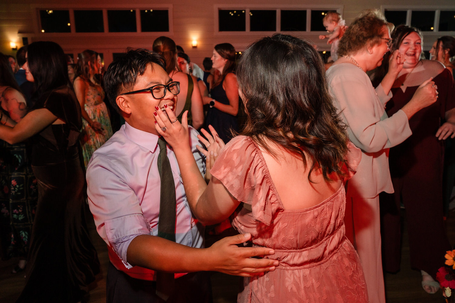 A person in a dress and a person in a dress shirt and tie laugh together on a dance floor at a party.
