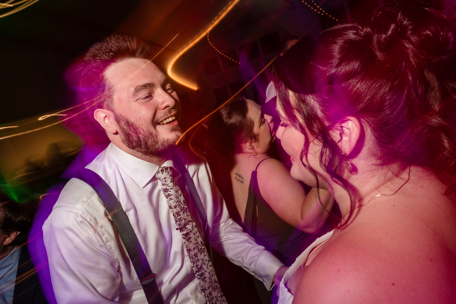 A smiling man in a dress shirt and suspenders dances with a woman at a party with long-exposure light trails.