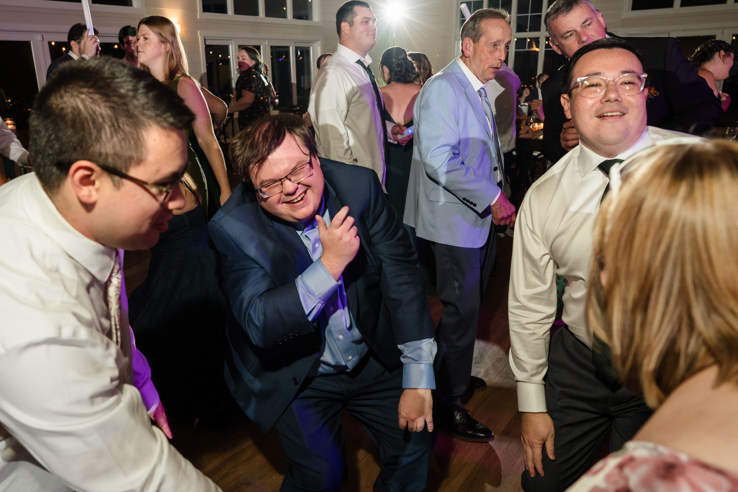 People dancing at an indoor event, with a man in a navy suit smiling as he dances in the center of the frame.